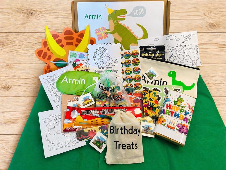 May include: A dinosaur-themed birthday gift set with various items. Includes a cardboard box, a green eye mask, a canvas bag with "Armin" and a dinosaur graphic, stickers, coloring pages, and a birthday-themed cup. The box has a dinosaur illustration and the name "Armin".