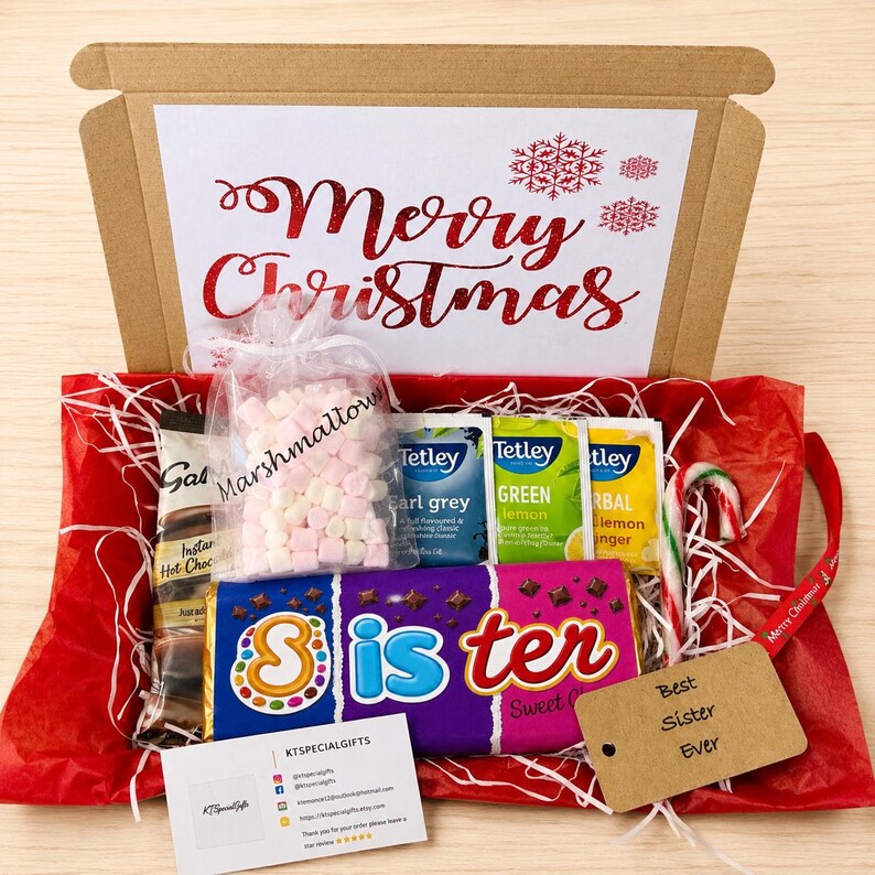 May include: A cardboard gift box with a Merry Christmas sign, containing a bag of marshmallows, Tetley tea bags, a candy cane, and a purple chocolate bar labeled Sister. A brown gift tag reads Best Sister Ever, and a business card is included.