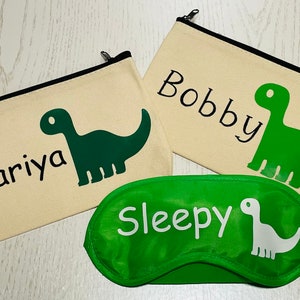 May include: Three items: two beige zippered pouches with black trim, each featuring a green dinosaur graphic and a name, and a green sleep mask with a white dinosaur and the word "Sleepy".