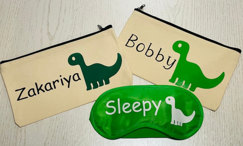May include: Three items: two beige zippered pouches with black trim, each featuring a green dinosaur graphic and a name, and a green sleep mask with a white dinosaur and the word "Sleepy".