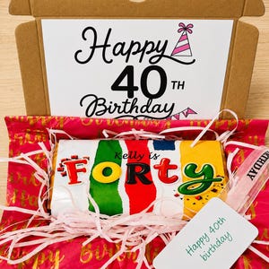 May include: A cardboard gift box containing a chocolate bar wrapped in colorful paper with the text Kelly is FORTY. The box lid features the text Happy 40TH Birthday with a party hat icon. Pink shredded paper filler and a Happy 40th birthday tag.
