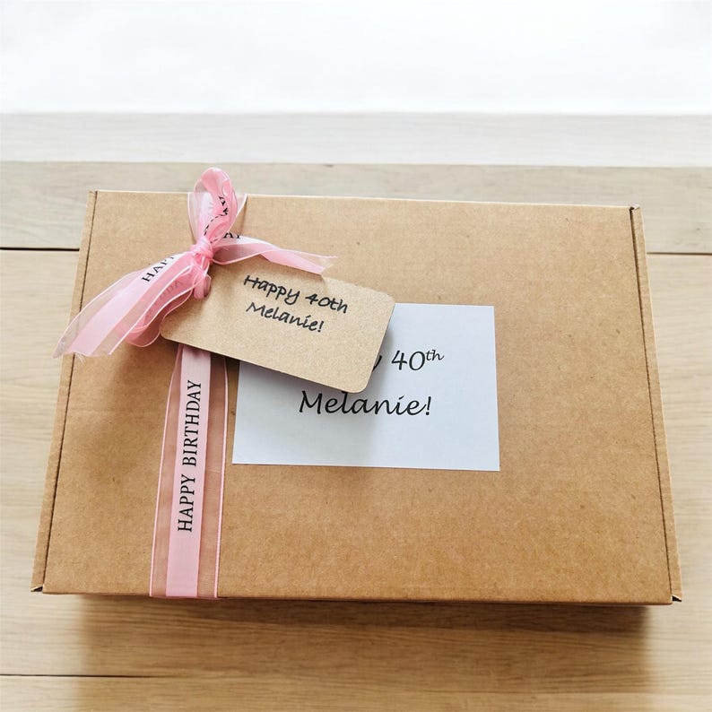 May include: A rectangular brown cardboard gift box decorated with a light pink ribbon printed with the words HAPPY BIRTHDAY. A tan gift tag reads Happy 40th Melanie! and a white rectangular label features the text 40th Melanie! in black font.