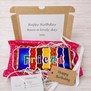May include: A gift box with a chocolate bar that says "Friend" in colorful letters. The box is lined with pink tissue paper and white ribbon. A tag says "Happy Birthday". The box also contains a card that says "Happy Birthday, Have a lovely day".
