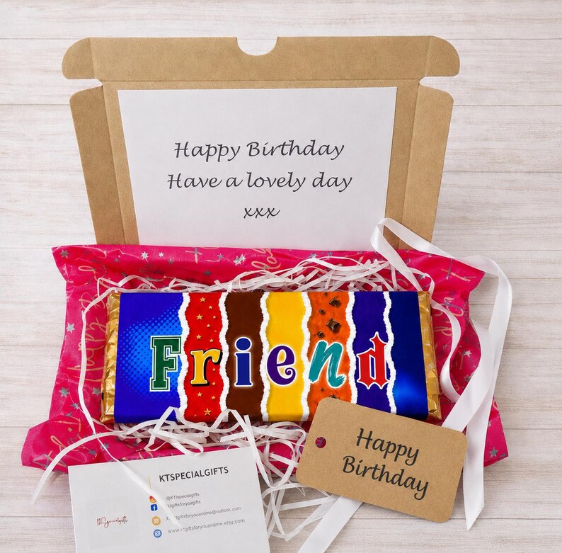 May include: A gift box with a chocolate bar that says "Friend" in colorful letters. The box is lined with pink tissue paper and white ribbon. A tag says "Happy Birthday". The box also contains a card that says "Happy Birthday, Have a lovely day".