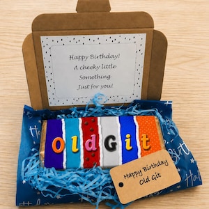 May include: A birthday gift box containing a chocolate bar with the words "old Git" in colorful letters. The box is brown and has a tag that says "Happy Birthday Old Git". Inside the box is a message that says "Happy Birthday! A cheeky little Something Just for you!"