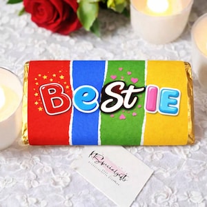 May include: A colorful candy bar wrapper with the word "Bestie" in a playful font. The wrapper is divided into red, blue, green, and yellow sections. Small hearts and stars decorate the design. A small white card with text is in the foreground.
