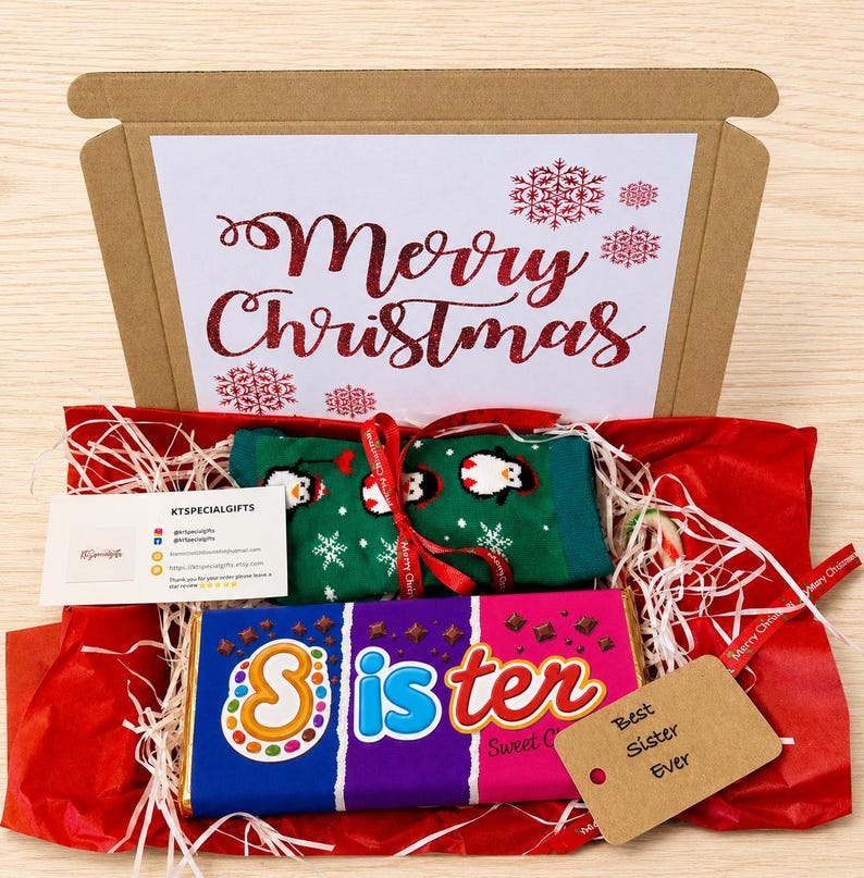 May include: A gift box containing green socks with a penguin pattern, a chocolate bar with a wrapper reading Sister, a white card with Merry Christmas in red glitter text, a small business card, and a brown tag labeled Best Sister Ever.