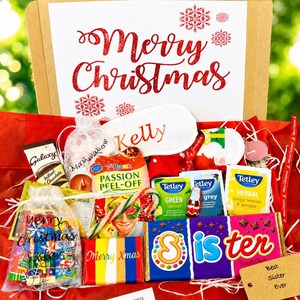 May include: A gift box containing a Merry Christmas sign, a white sleep mask labeled Kelly, a Galaxy hot chocolate packet, a 7th Heaven face mask, Tetley tea bags, a candy cane, a chocolate bar labeled Sister, and a tag reading Best Sister Ever.