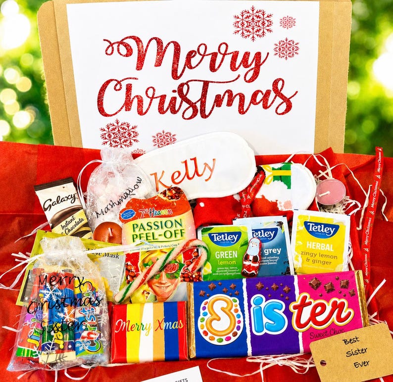 May include: A gift box containing a Merry Christmas sign, a white sleep mask labeled Kelly, a Galaxy hot chocolate packet, a 7th Heaven face mask, Tetley tea bags, a candy cane, a chocolate bar labeled Sister, and a tag reading Best Sister Ever.