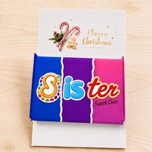 May include: A rectangular chocolate bar wrapper featuring a blue, purple, and pink gradient background with torn paper edges. The word Sister is printed in large, colorful, stylized letters, with Sweet Choc text below. A Merry Christmas card is attached.