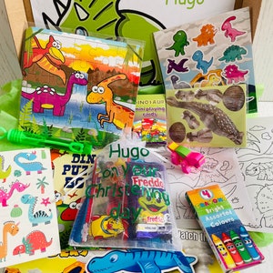 May include: A gift box filled with dinosaur-themed toys and treats. Includes a puzzle, stickers, crayons, and a bag of candy. The box is decorated with a green dinosaur head and the name "Hugo".