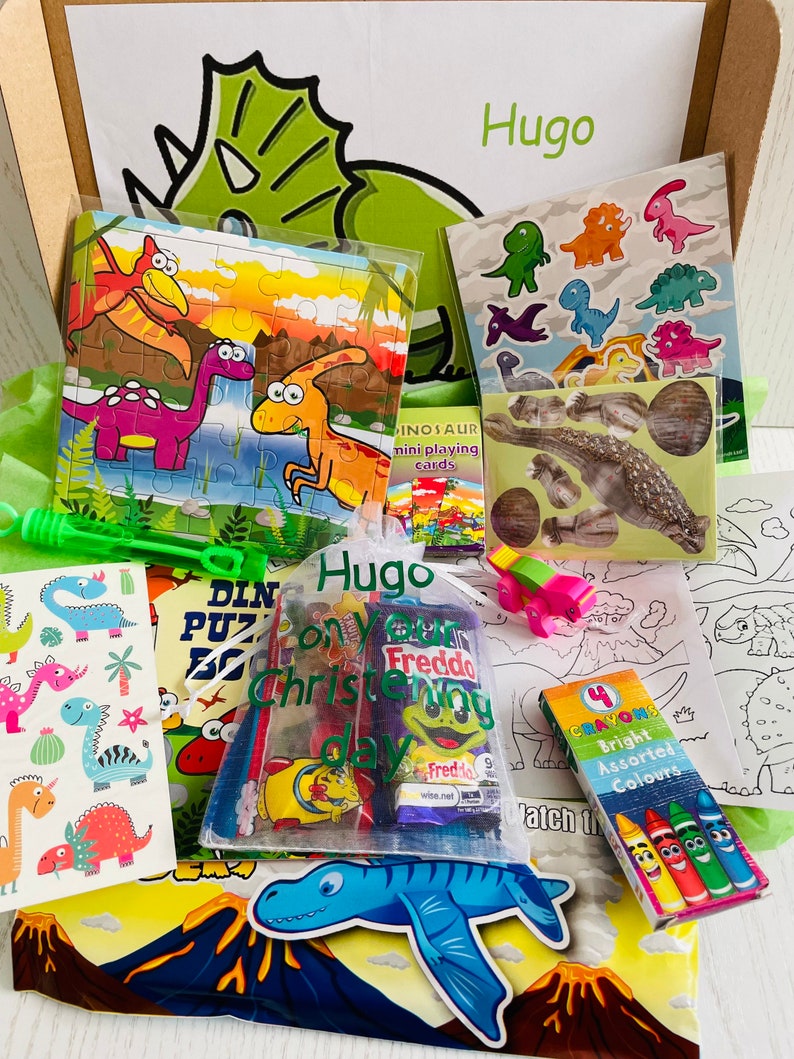May include: A gift box filled with dinosaur-themed toys and treats. Includes a puzzle, stickers, crayons, and a bag of candy. The box is decorated with a green dinosaur head and the name "Hugo".