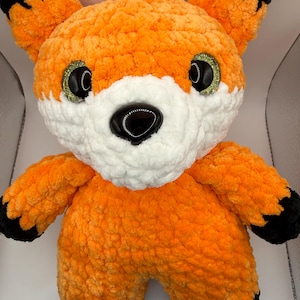 May include: A plush fox toy crafted from soft orange, white, and black yarn. The fox features large, glossy black eyes with green glitter accents, a smooth black plastic nose, and pointed ears with black tips. The body is round and textured.