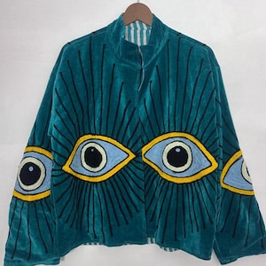 May include: Teal velvet jacket with embroidered eye designs. The jacket features a high collar and a striped lining. The eye designs are in blue, yellow, and black. The jacket is hanging on a wooden hanger.