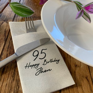 Könnte beinhalten: Eine weiße Serviette mit dem Text "95 Happy Birthday Jhon" in schwarzer Tinte. Eine silberne Gabel und ein Messer liegen auf der Serviette.