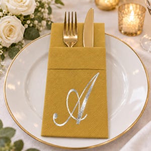Puede incluir: Una servilleta de papel color amarillo mostaza doblada en forma de bolsillo rectangular, con una letra A cursiva en plata metálica impresa en el frente. Un tenedor y un cuchillo de metal dorado están insertados dentro del bolsillo de la servilleta.
