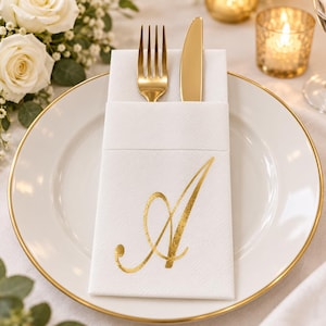 Puede incluir: Una servilleta de papel blanca doblada en forma de bolsillo, con la letra A en caligrafía dorada. La servilleta está colocada sobre un plato blanco con borde dorado, conteniendo un tenedor y un cuchillo dorados. La textura es visible.