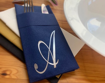 May include: A navy blue napkin with a silver letter A monogram. The napkin is folded in half and has a pocket for silverware. The napkin is on a wooden table.