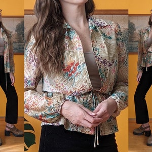 May include: A vintage floral print blouse with a tie waist. The blouse features a beige background with colorful floral patterns in red, purple, and green. The sleeves are long and gathered at the wrists. The blouse is paired with black pants.