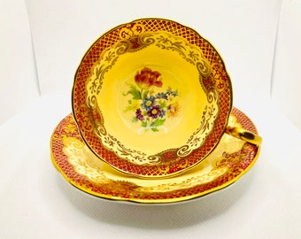 May include: A vintage teacup and saucer set with a yellow background and gold trim. The cup features a floral design with pink, purple, and blue flowers. The saucer has a red and gold pattern.