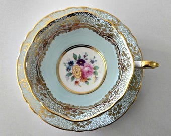 May include: A porcelain teacup and saucer set featuring a pale blue interior and a white centre medallion with a colourful floral bouquet. The edges are decorated with intricate gold filigree and a repeating geometric pattern in gold and white.