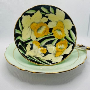 May include: A teacup and saucer set featuring a floral design. The teacup has a black interior with yellow and white daffodils. The saucer and the cup's exterior are light green with gold trim. The handle is white with gold accents.