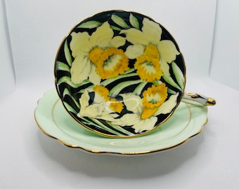 May include: A teacup and saucer set featuring a floral design. The teacup has a black interior with yellow and white daffodils. The saucer and the cup's exterior are light green with gold trim. The handle is white with gold accents.