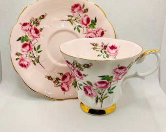 May include: A pink teacup and saucer set with a floral pattern of pink roses and green leaves. The set has a gold trim around the edges.