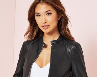 Pode incluir: Casaco de couro preto com gola alta e zíper prateado. O casaco tem um corte sob medida e apresenta detalhes sutis de costura nos ombros. A modelo veste um top branco e jeans pretos.