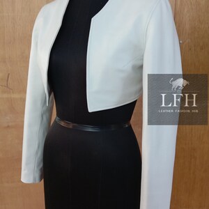 May include: A white leather cropped jacket with long sleeves. The jacket has a rounded neckline and a slightly fitted silhouette.