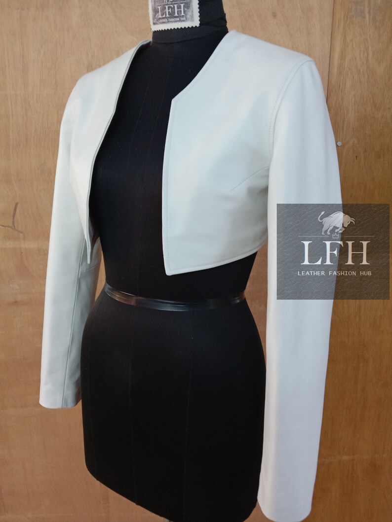 May include: A white leather cropped jacket with long sleeves. The jacket has a rounded neckline and a slightly fitted silhouette.