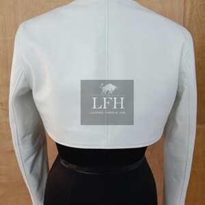 May include: A white cropped leather jacket with long sleeves. The jacket has a simple design and is made of soft, supple leather. The jacket is shown on a mannequin with a black skirt.