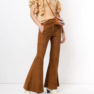 Pode incluir: Uma mulher est&aacute; vestindo uma blusa bege com babados, uma cal&ccedil;a flare de camur&ccedil;a marrom e botas de salto branco. Ela est&aacute; carregando uma pequena bolsa marrom.