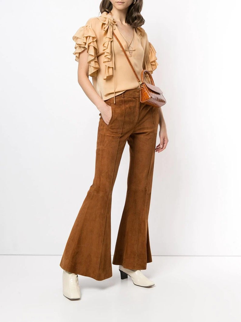Pode incluir: Uma mulher est&aacute; vestindo uma blusa bege com babados, uma cal&ccedil;a flare de camur&ccedil;a marrom e botas de salto branco. Ela est&aacute; carregando uma pequena bolsa marrom.
