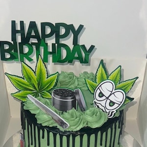 May include: A black and green cake with green frosting and a marijuana leaf design. The cake is decorated with a "Happy Birthday" topper and a marijuana leaf with a cartoon face.