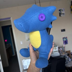 Puede incluir: Un peluche de caballito de mar azul hecho a mano con una panza amarilla y un botón morado como ojo. El peluche está hecho de tela suave y tiene un diseño caprichoso, perfecto para niños o como objeto decorativo.