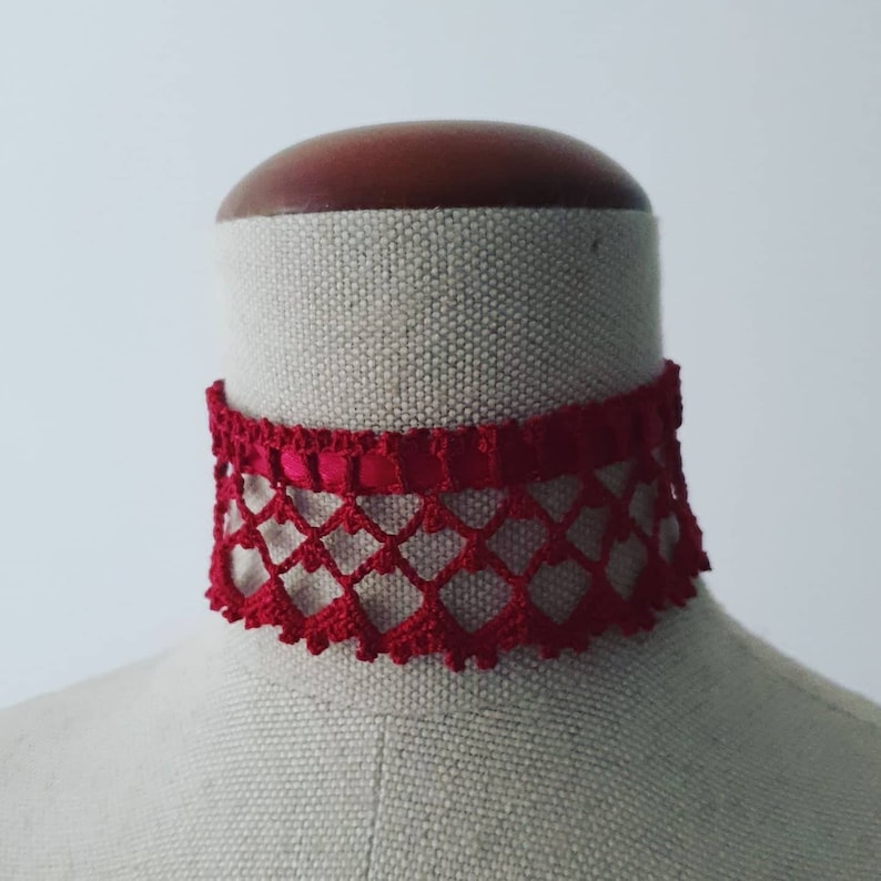 May include: A red crocheted choker necklace with a heart-shaped design. The necklace is on a fabric-covered mannequin.
