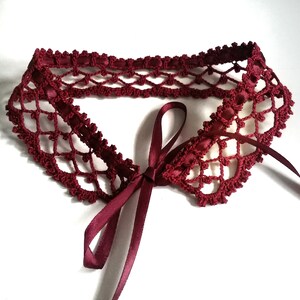 May include: A burgundy crocheted collar with a matching satin ribbon tie. The collar is made with a delicate openwork pattern.