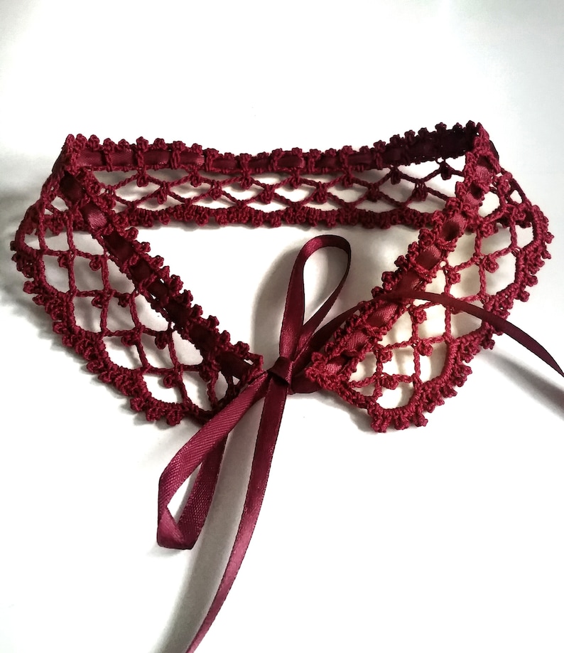 May include: A burgundy crocheted collar with a matching satin ribbon tie. The collar is made with a delicate openwork pattern.