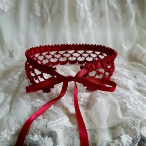May include: A red crocheted lace collar with a matching ribbon tied in a bow. The collar is delicate and intricate, with a delicate pattern.