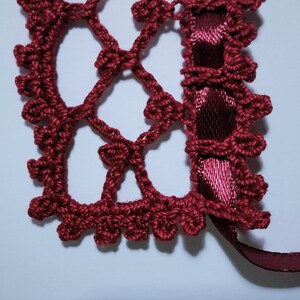 May include: A close-up of a burgundy crocheted lace trim with a burgundy satin ribbon attached. The lace is made of small, delicate stitches and has a delicate, feminine look.