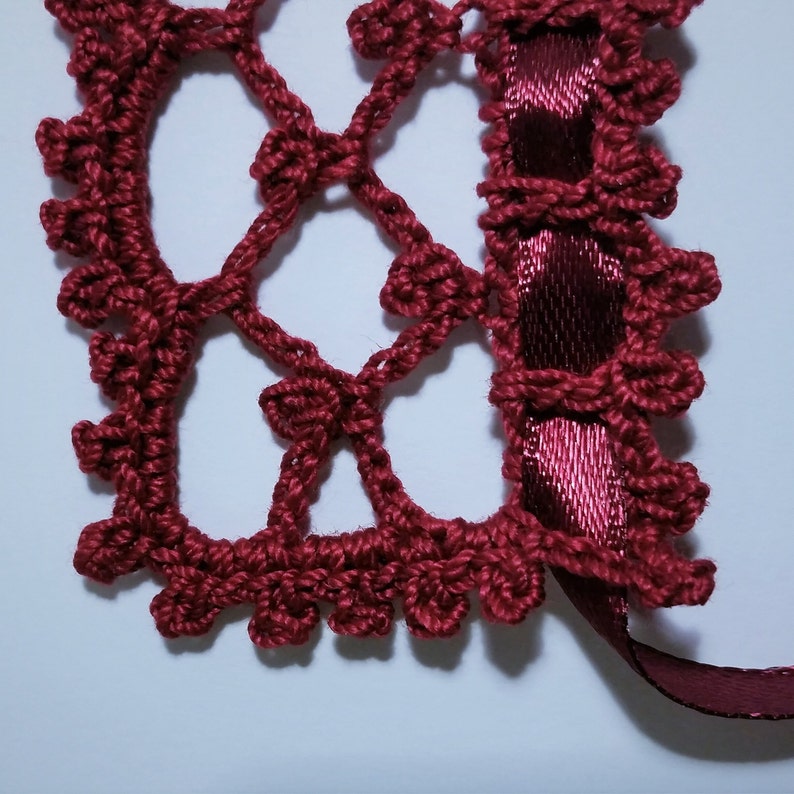May include: A close-up of a burgundy crocheted lace trim with a burgundy satin ribbon attached. The lace is made of small, delicate stitches and has a delicate, feminine look.