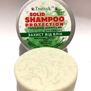 Puede incluir: Una barra de champú sólida redonda y blanca en una lata de plata con el texto "Tsutsyk Solid Shampoo Protection" en letras verdes y negras. La lata también tiene el texto "твердий шампунь для тварин" en letras negras y "Вага нетто 75г" en letras negras. La barra de champú está diseñada para proteger a las mascotas de las pulgas.