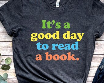 Puede incluir: Una camiseta gris oscuro con el texto "It's a good day to read a book." en letras de colores. La camiseta se combina con jeans, un sombrero y zapatos.