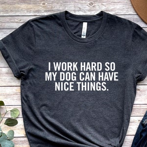 May include: A dark grey t-shirt with the text "I work hard so my dog can have nice things." printed in white. The shirt is shown on a wooden background with a pair of brown sandals, blue jeans, and a brown hat.