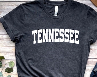 Puede incluir: Una camiseta gris oscuro con la palabra "TENNESSEE" impresa en blanco en la parte delantera. La camiseta se combina con pantalones vaqueros azules, sandalias marrones y un sombrero marrón.
