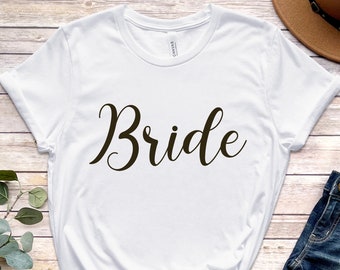 Puede incluir: Camiseta blanca con la palabra "Bride" escrita en cursiva negra. La camiseta está sobre una superficie de madera con un par de sandalias marrones, unos vaqueros azules y un sombrero marrón.