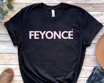 Può includere: Maglietta nera con una grafica rosa che dice "FEYONCÉ" su uno sfondo di legno. La maglietta è abbinata a un cappello marrone, jeans blu e sandali marroni.