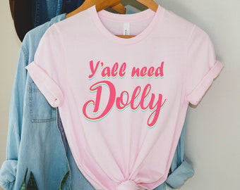 May include: Pink t-shirt with the text "Y'all need Dolly" in a rainbow-colored font.