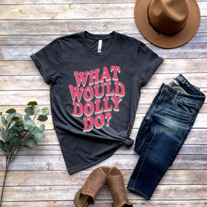May include: A dark gray t-shirt with the text "What Would Dolly Do?" in a retro, rainbow-colored font. The shirt is laid flat on a wooden surface with a pair of brown sandals, blue jeans, and a brown hat.
